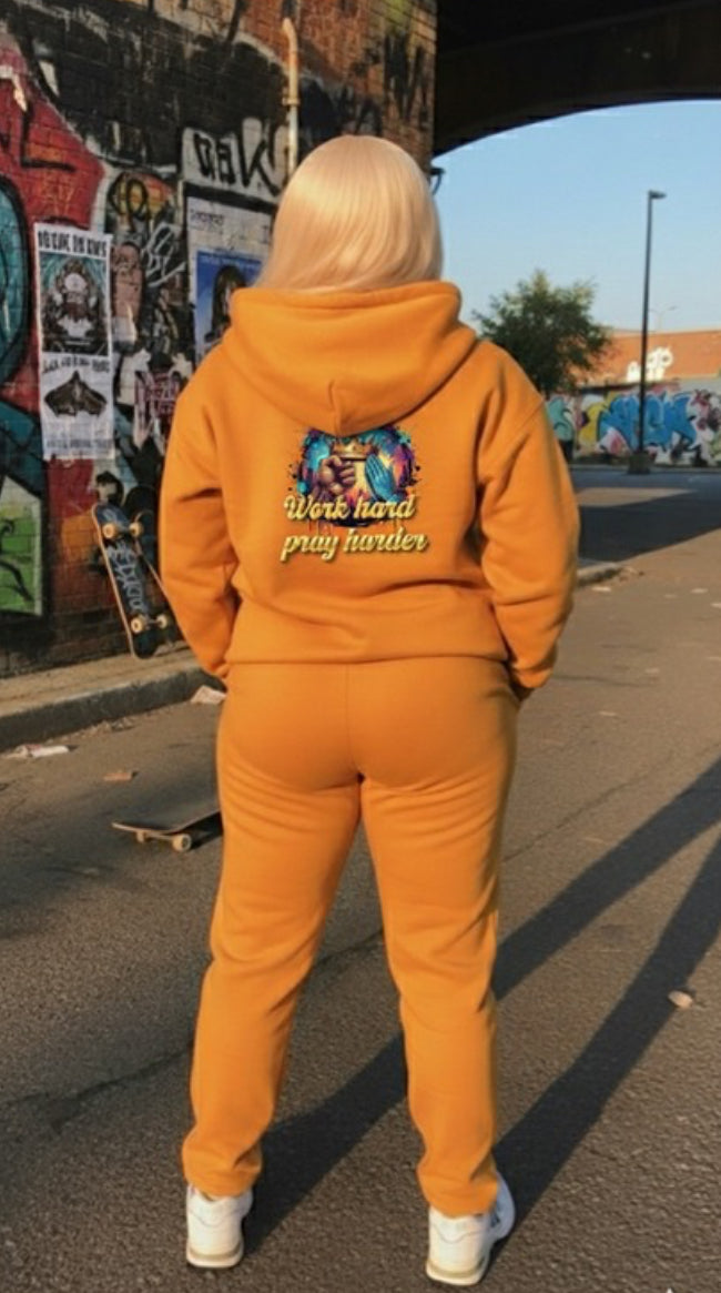 A person wearing a Work Hard- HOODIE in orange, showcasing streetwear style in an urban setting.